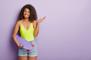 Extreme sport and adventure concept. Positive African American woman holds skateboard, raises palm and smiles pleasantly, wears casual outfit isolated on purple wall. Skater girl with longboard indoor