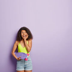 Happy dark skinned female enjoys extreme riding on skateboard, has curly hair, keeps hand on cheek, looks with dreamy expression, stands over purple wall. Skater style. Lovely woman with penny board