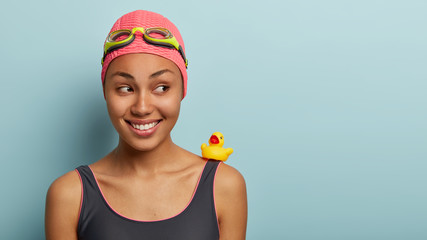 Close up shot of smiling charming good looking swimmer feels relaxed and delighted after swimming in pool, wears swimhat with goggles, carries small yellow toy duckling on right shoulder, looks aside
