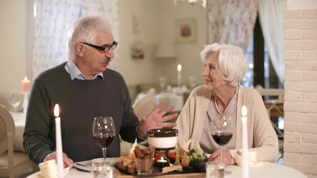 Waist-up shot of 60-something senior Caucasian gentleman dining with his wife in candlelit restaurant, telling her something and gesturing animatedly while she is listening and offering her opinion