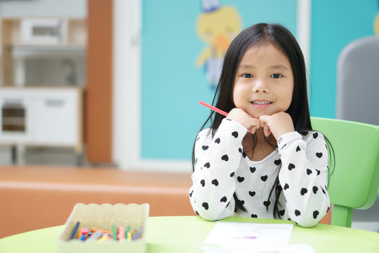 Asian child or kid girl and student happy smiling and learning for coloring or paint on white paper with teacher at kindergarten classroom and nursery or pre school on colorful table and background