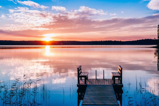 Idyllic view of the long pier with wooden bench on the lake. Sunset or sunrise over the water.
