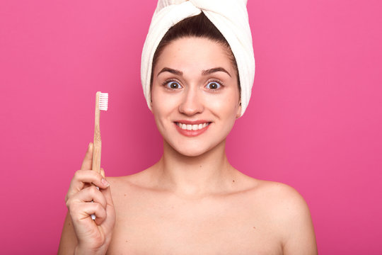 Portrait of smiling pretty woman with astonished facial expression, holds toothbrush isolated over pink background and looking directly at camera, posing with nacked shoulders and white towel on head.