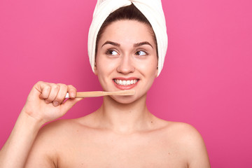 Young cheerful woman with perfect white smile looking aside, holding wooden toothbrush in front of face, being naked, having smooth well kept skin, posing isolated over pink background in studio.