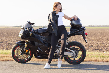 Fototapeta na wymiar Full length portrait of attractive good looking female looking aside, standing near her vehicle, putting her arm on helmet, wearing leather jacket, white t shirt, black trousers and white sneakers.