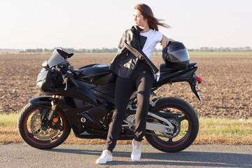 Fototapeta na wymiar Adorable magnetic biker having trip on her motorcycle, standing near field, enjoying splendid views, looking aside, being in good mood, having confident facial expession. Extereme sports concept.