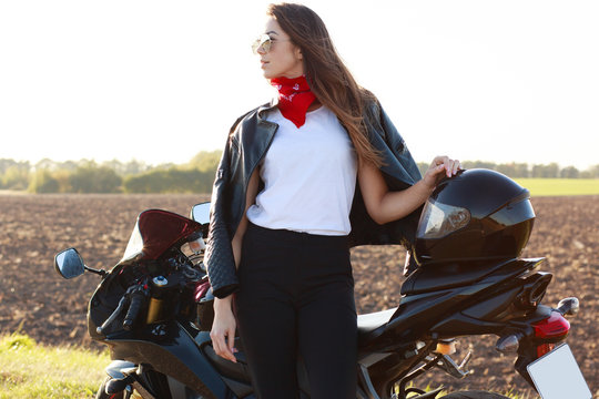 Outdoor Shot Of Confident Risky Young Woman Standing Near Her Motorbike, Putting Hand On Helmet, Wearing Red Bandana, White Shirt, Leather Jacket And Black Trousers. Motorcycle Tourism Concept.