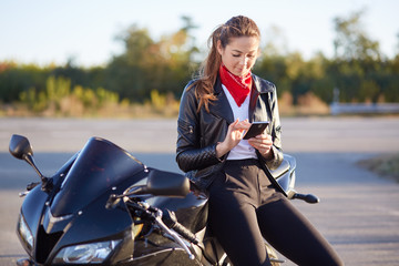 Fototapeta na wymiar Young female biker with phone in hands, typing message on cellular, wears red bandana, white shirt and leather jacket, poses on motorbike. Technology and lifestyle, high speed, traveling on motorcycle