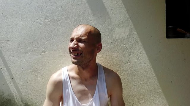 Young positive skinny anorexic bald happy smiling homeless man sitting on the urban street in the city or town near white wall with big smile looking at the camera, homelessness social documentary