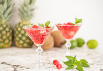 Two refreshing red translucent drinks with ice and raspberries. The raspberry juice is garnished with a green leaf, and there are pieces of ice and raspberries scattered around the drinks