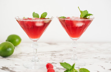 Two refreshing red translucent drinks with ice and raspberries. The raspberry juice is garnished with a green leaf, and there are pieces of ice and raspberries scattered around the drinks