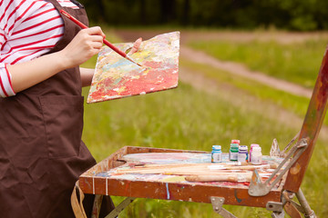 Outdoor shot of unknown woman around nature, wearing brown apron, holding brush and palette, making sweeping brushstroke, creating open air sketches, tubes with paints are on view. Art concept.