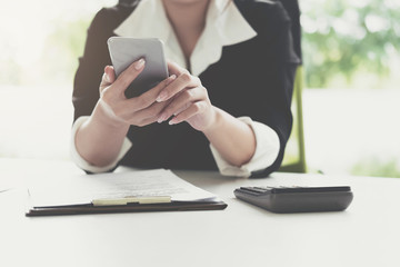 Businesswoman in black suit thinking about his project, using smart phone looking at the screen, working on paper with calculator, business data, marketing report in office
