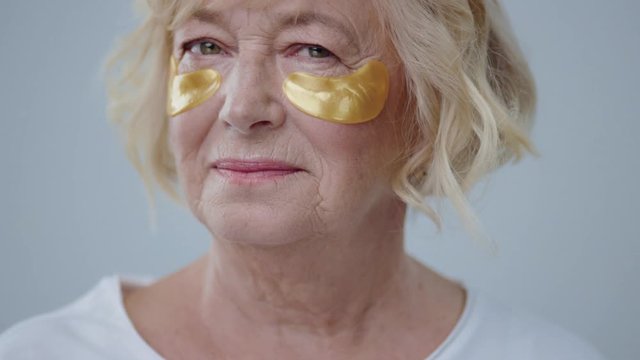 Appealing blonde older woman with blonde hair and golden eye patches on face looking at camera posing in the photo studio against the wall.