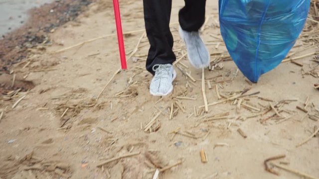 Closeup tilt up shot of unrecognizable volunteer with trash picker and bag walking along coastline and collecting waste on dirty beach