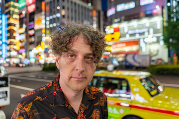 A middle aged man enjoys the nightlife in Shinjuku in Tokyo, Japan, with the beautiful city lights and traffic behind.