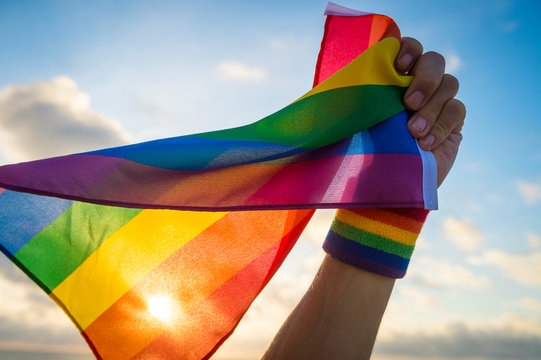 Hand with rainbow wristband waving gay pride flag waving backlit in the wind against golden sunset sky