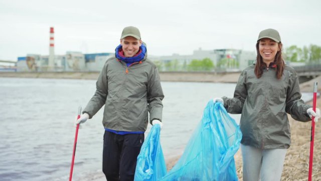 Two smiling volunteers, young man and woman, talking while carrying and tying trash bags after cleaning beach, tilt up and down shot