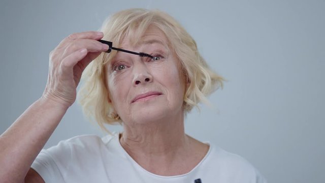 Close-up Of Gorgeous Senior Woman With Blonde Hair Applying Mascara On Eyelashes. Pretty Older Woman Doing Make-up Posing In The Studio.