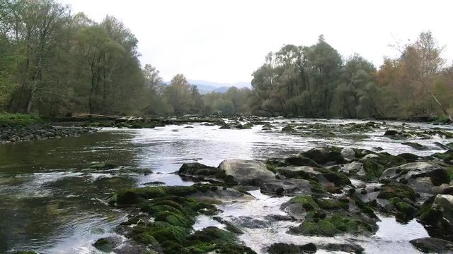 Drone moving above a river rippling onto rocks surrounded by lush forest