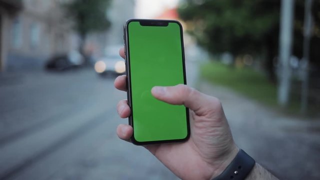 Sociable guy using a modern smartphone with chromekey greenscreen outdoors. Close-up of male hand holding a mockup device on blurred background.