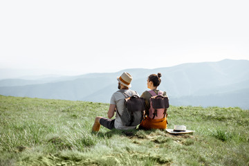 Young couple sitting on the green meadow, traveling with backpacks in the mountains during the...