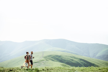 Fototapeta premium Couple walking with backpacks on the green meadow, traveling in the mountains during the summer time, wide landscape view