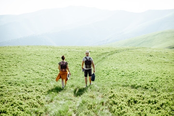 Couple walking with backpacks on the green meadow, traveling in the mountains during the summer time, wide landscape view