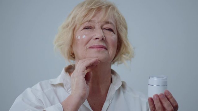 Portrait Of Cheerful Older Woman Applying Moisturizer To Her Face Looking In The Mirror. Wonderful Senior Model With Blonde Hair In White Shirt Holding Cream Isolated On Grey Background.