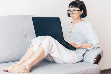 Fototapeta na wymiar Middle-aged brunette woman in glasses on the gray sofa using laptop .