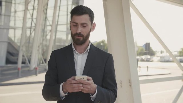 Closeup Portrait of Smiled Handsome Businessman Texting Massage on his Phone. Successful Man standing near the Modern Glassy Business Building in classical Suit. Charmingly Smile. Social Network.