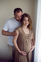 pregnant girl in a brown dress is standing at the window with her husband