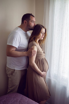 pregnant girl in a brown dress is standing at the window with her husband