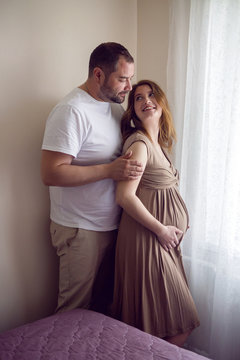 pregnant girl in a brown dress is standing at the window with her husband