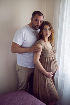 pregnant girl in a brown dress is standing at the window with her husband