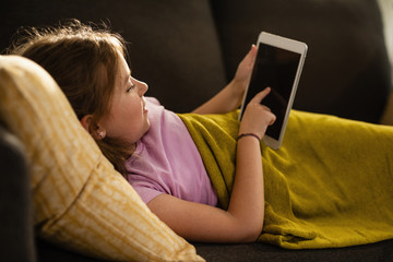 Little girl playing games on touchpad while relaxing on the sofa.