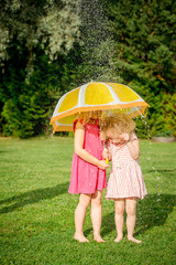 Two young girls sisters standing outdoors in garden under umbrella with water drops falling, sisters smiling and having fun. Childhood memories concept.