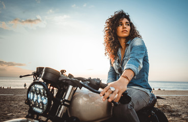 Schönes Mädchen, das Spaß daran hat, ihr individuelles Cafe-Racer-Motorrad zu fahren und den Sonnenuntergang am Strand zu genießen