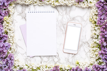 Mobile phone mock up, notebook and frame of white and lilac flowers on marble table in flat lay style.