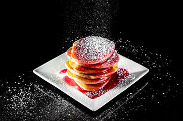 Powdered sugar sprinkles to stack of pancaces on black reflective background.