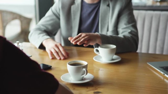 Closeup of two anonymous businessmen talking over coffee during business meeting in cafe, tracking left closeup shot. Open laptop, coffee cups, notepad with pen and smartphone on table.
