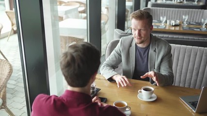 High angle view tracking right shot of two men having business meeting over coffee and tea in cafe. Middle aged businessman talking to unrecognizable colleague, open laptop on table
