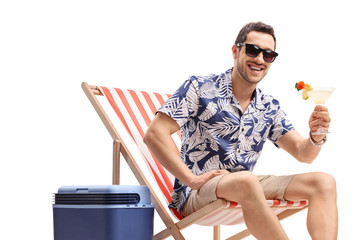 Young guy with a cocktail sitting in a deck chair next to a cooling box and looking at the camera