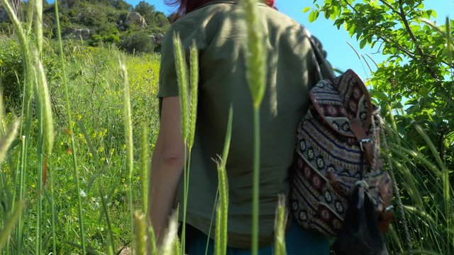 A young red-haired traveler girl in a cowboy hat walks along a mountain path overgrown with grass and shrubs with yellow flowers.