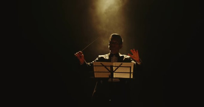 Unrecognizable male symphony orchestra conductor wearing black tux is directing musicians in orchestra pit by moving his hands and baton, studio shot on black background 4k footage