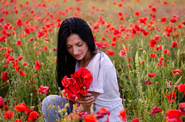 Naklejka premium a young girl with dark long hair is wearing a blouse T-shirt and blue jeans sitting with a bouquet of poppy flowers among the poppy field