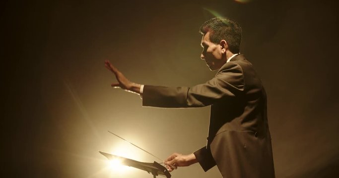 Asian male orchesra conductor wearing tux is directing symphony orchestra with movement of his hands and stick, studio shot on black background 4k footage