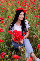 A young girl with dark long hair in a white T-shirt and blue jeans sits among poppy field