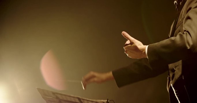 Unrecognizable male symphony orchestra conductor wearing black tux is directing musicians in orchestra pit by moving his hands and baton, studio shot on black background 4k footage