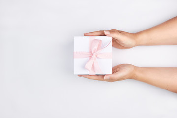 female hands holding white gift box with pink ribbon on white background flatlay scene top view